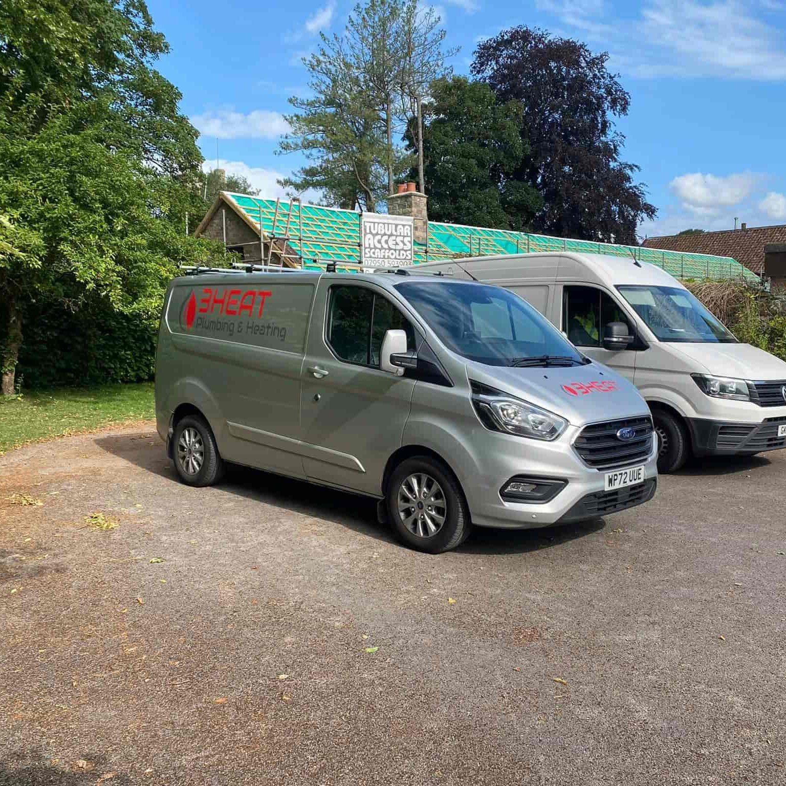 A silver van with "3HEAT Plumbing & Heating" branding is parked on a paved area beside another van, surrounded by trees and a house with scaffolding in the background.
