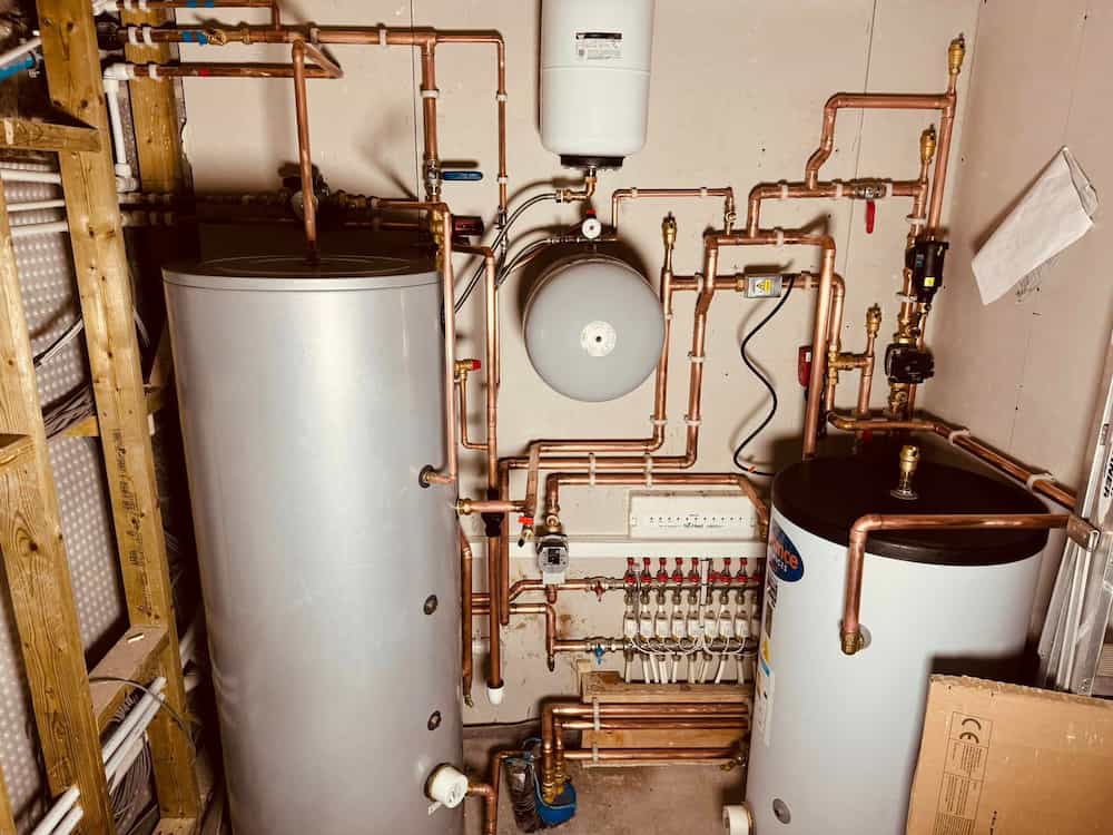 A utility room with water heaters, a hot water cylinder, an expansion tank, copper pipes, valves, gauges, and a central heating manifold mounted on the wall.