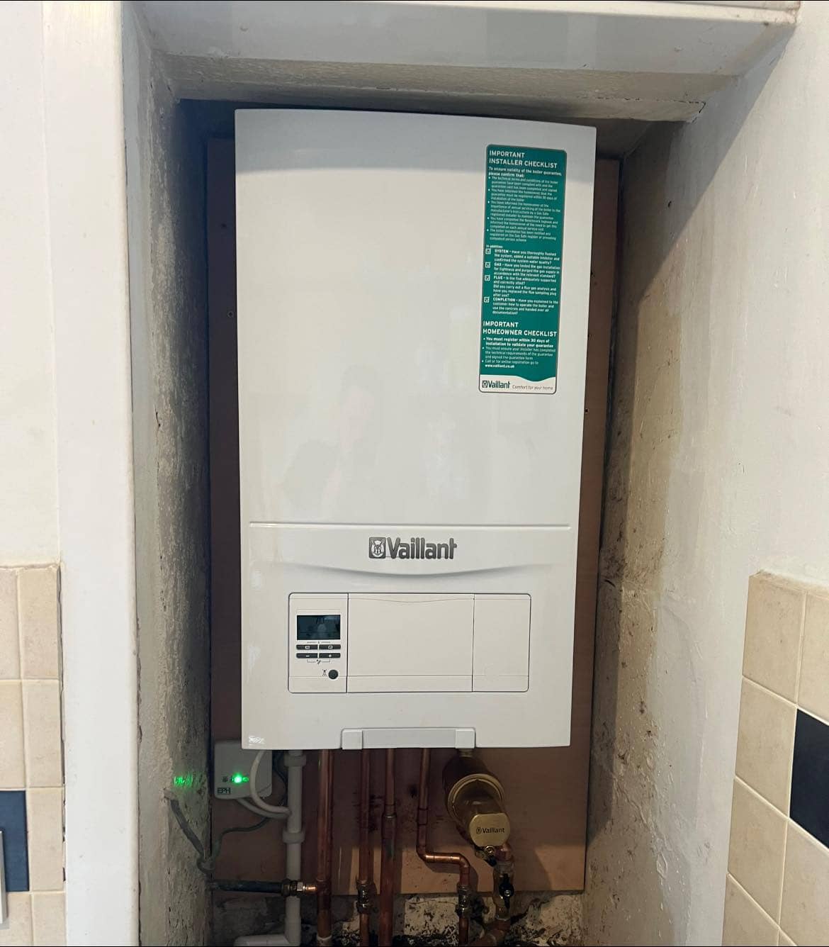 A wall-mounted Vaillant boiler is installed in a recessed space, with visible pipes and wires connected below. The boiler has a green sticker on the upper right and is surrounded by a tiled and painted wall.