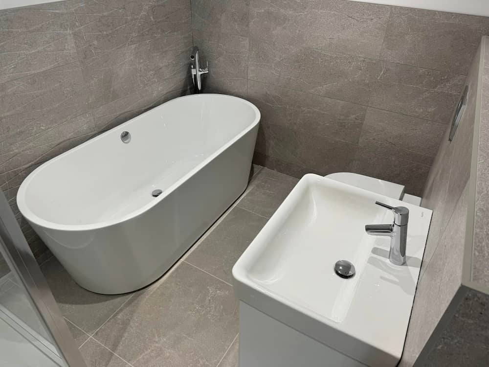 Modern bathroom with grey tiled walls and floor, featuring a white freestanding bathtub, a white rectangular sink with a silver faucet, and a partial view of a toilet beside the sink.