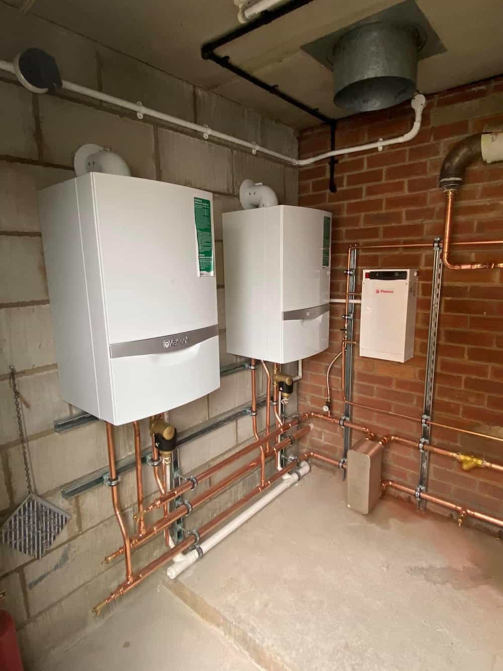 Two white wall-mounted boiler units with exposed copper and plastic pipes in a utility room with brick and concrete walls. A smaller white control box is mounted beside them, with ventilation ducting overhead.