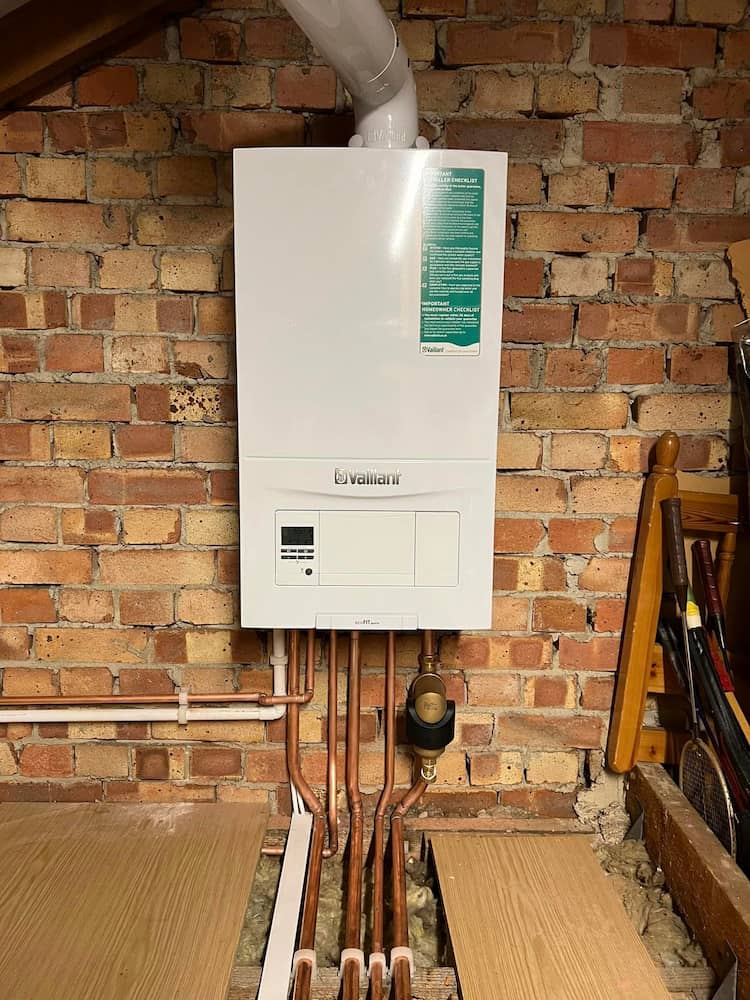 A white Vaillant boiler is mounted on a brick wall, with several copper pipes running vertically below it and floorboards partially lifted, revealing pipes beneath. Various tools and equipment are nearby.