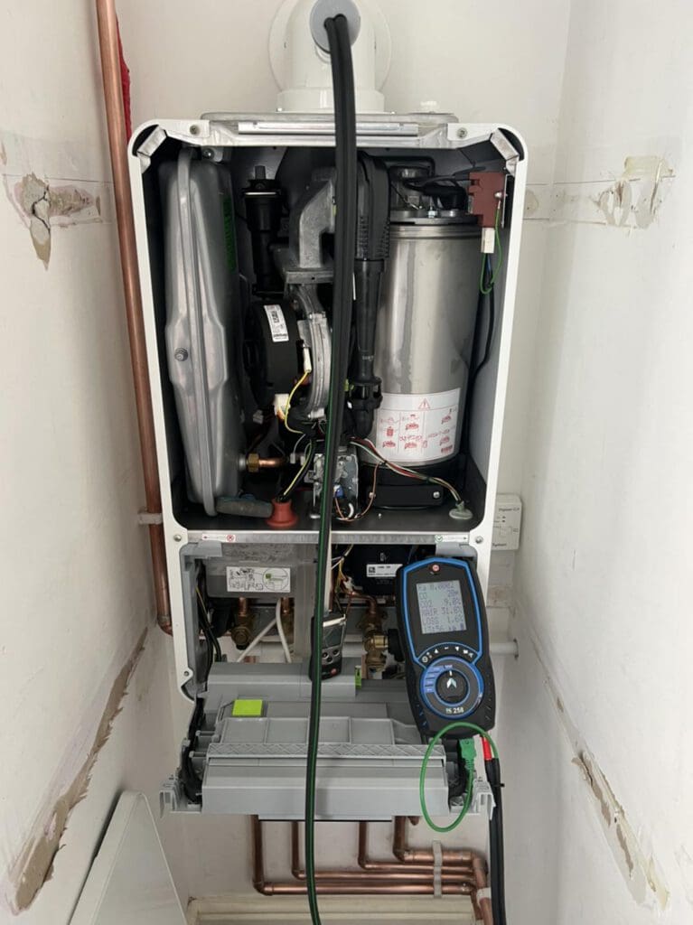 An open boiler unit with exposed internal components is being tested with a diagnostic device, which displays readings on its screen. Copper pipes and wiring are visible beneath and around the unit.