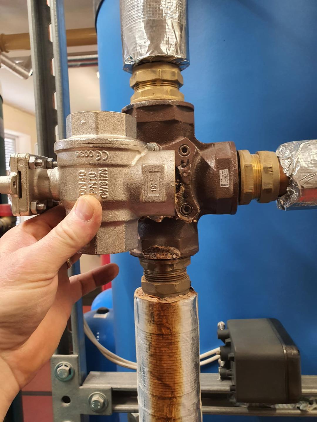 A person’s hand holds a metal valve connected to insulated pipes in an industrial setting, with a blue cylindrical tank and various fittings visible in the background.