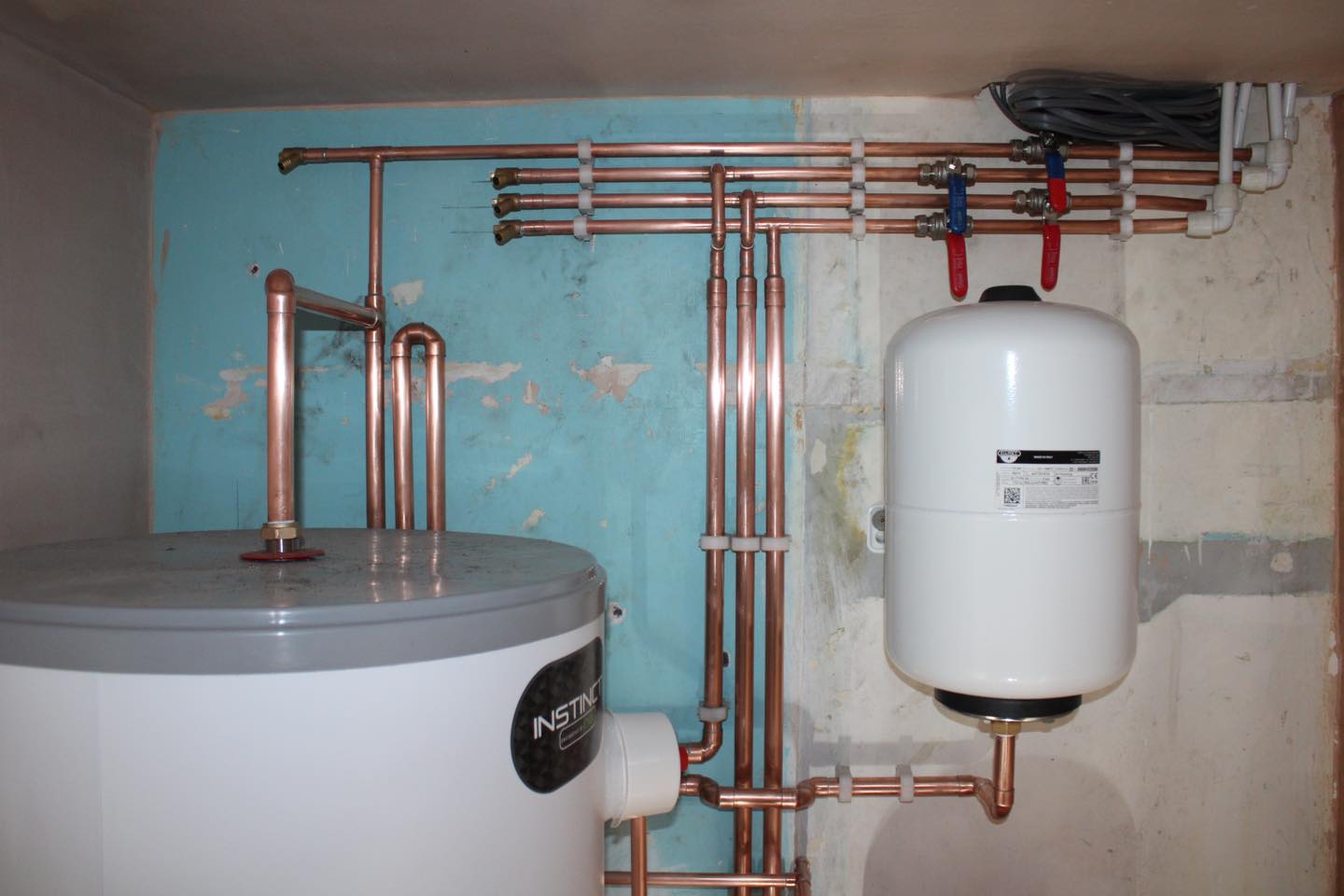 White water heater system with connected copper pipes on a blue and beige wall; a white expansion vessel is mounted above, with red and blue shut-off valves and electrical wires visible in the top right corner.