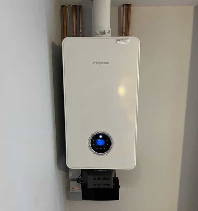 A white Worcester boiler is mounted on a wall, with copper pipes connected at the top and a digital display on the front. There are electrical outlets and cables below the unit.