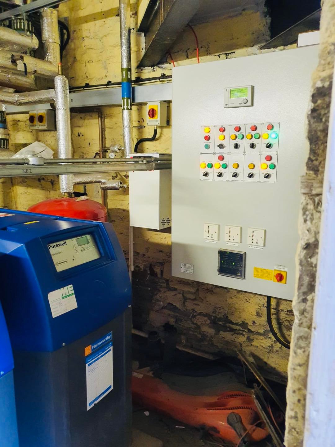 A utility room with exposed brick walls showing an electrical control panel with buttons and switches, pipes, gauges, and two blue industrial machines next to each other.