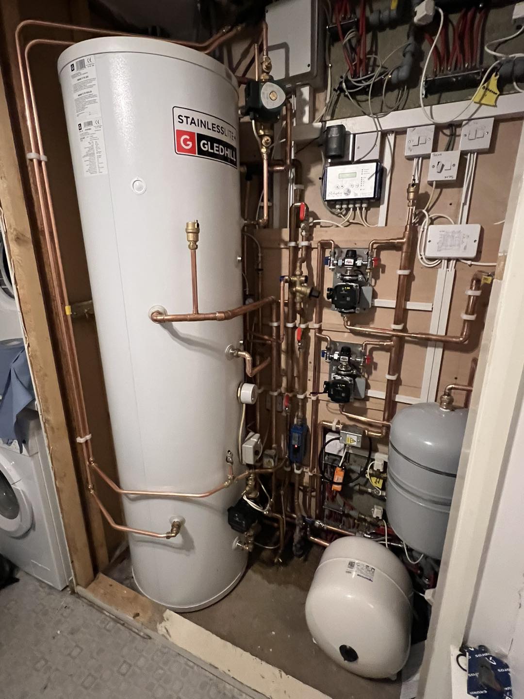 A hot water cylinder with copper pipes, gauges, and pumps installed neatly in a utility cupboard, surrounded by electrical wiring and control panels. A washing machine is partially visible on the left.
