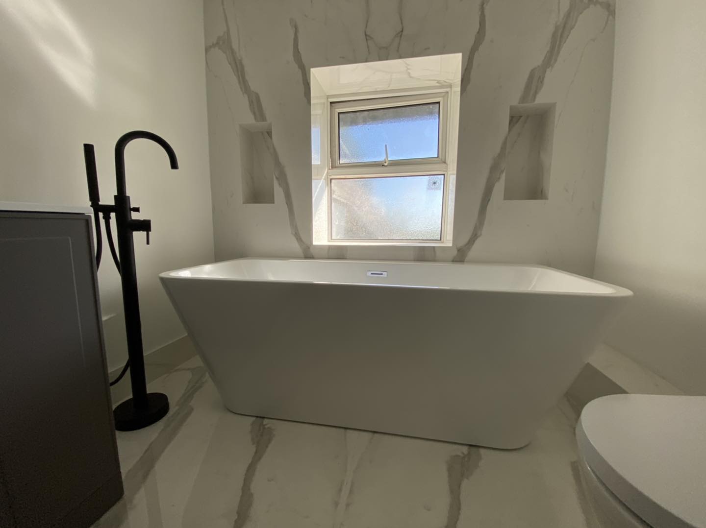 A modern bathroom with a white freestanding bathtub, a matte black floor-mounted faucet, marble walls, two recessed shelves, and a frosted window letting in sunlight.
