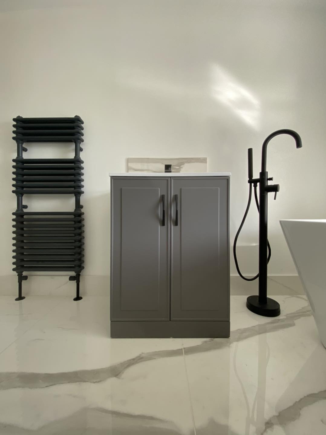 A modern bathroom features a gray vanity cabinet, a black towel rack, a freestanding black faucet, and part of a white bathtub on a white marble floor with gray veins.