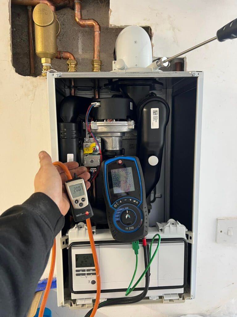 A person holds a digital multimeter and a temperature probe, testing a boiler with its front panel open. Visible wiring and copper pipes are connected above the unit.