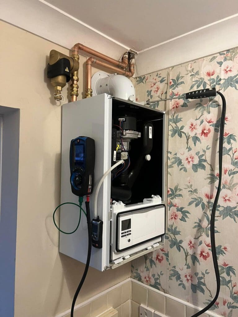 A wall-mounted boiler with its front panel open, showing internal components and connected diagnostic tools. Copper pipes and wires are attached above, next to a floral wallpapered wall and a beige painted wall.