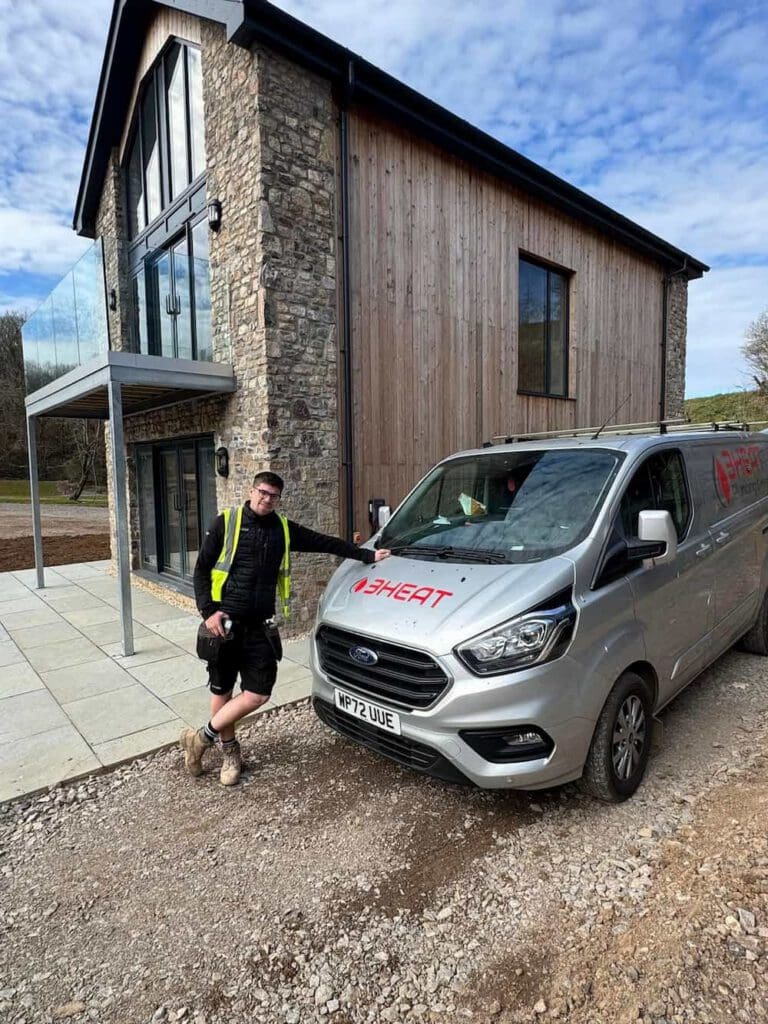 A man in workwear leans against a silver van with the "HEAT" logo, parked outside a modern house with stone and wood exterior, large windows, and a balcony on a clear day.