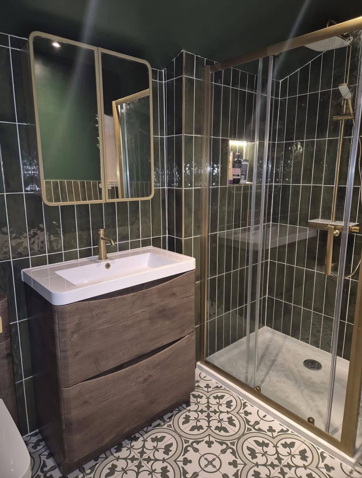 Modern bathroom with a green tiled shower, glass door, gold fixtures, a wooden vanity with white sink, large rectangular mirror, and patterned floor tiles in black and white.