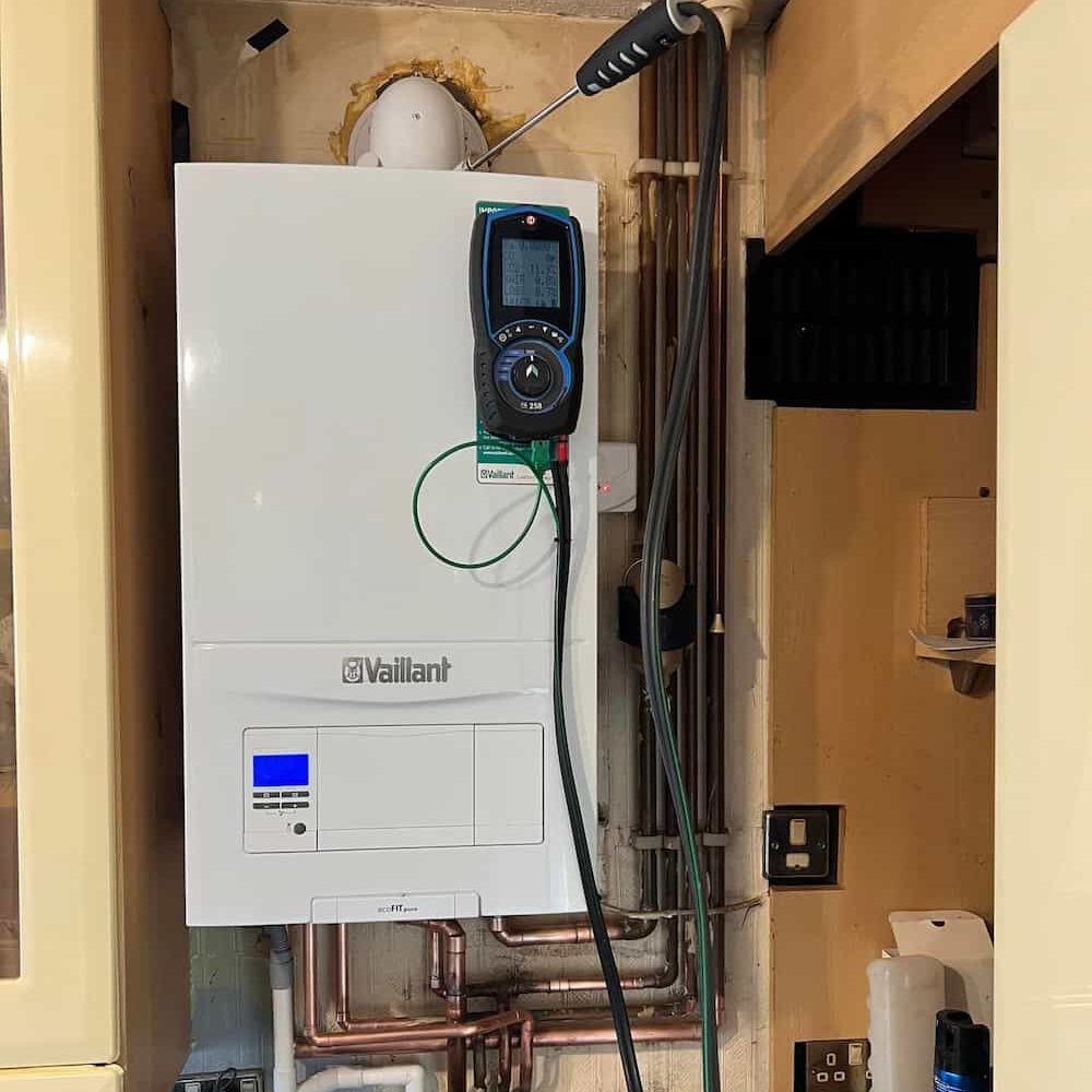 A white Vaillant boiler with a digital device attached hangs on a wall in a utility area. Pipes, wires, and a power outlet are visible around the boiler, and cabinets are on either side.