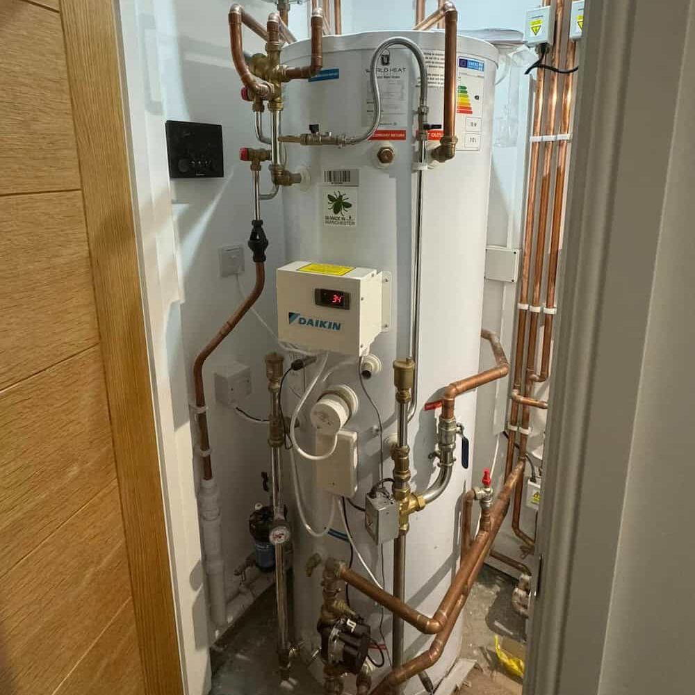 A hot water cylinder system with various copper pipes, gauges, valves, and a Daikin control unit installed in a small utility closet. The setup is neatly arranged and occupies most of the space.