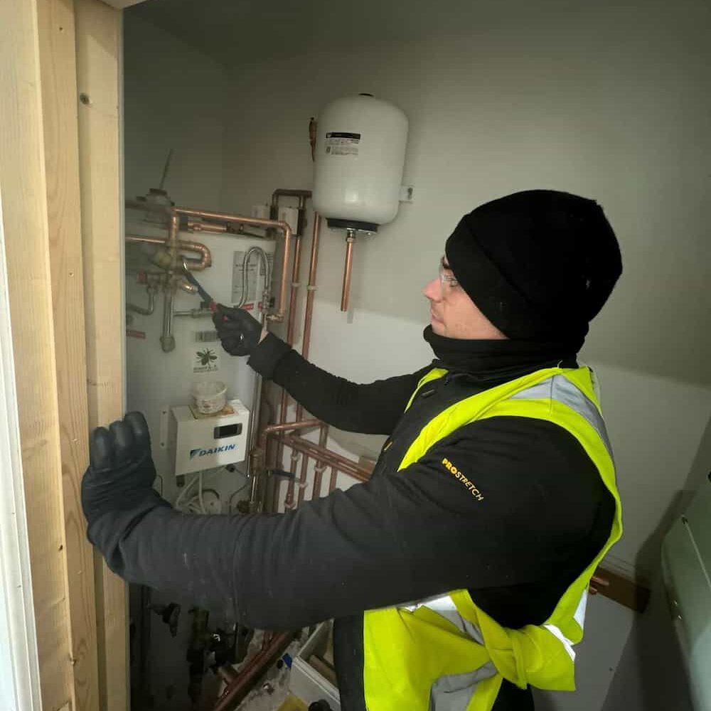 A person wearing a black beanie, gloves, and a yellow safety vest adjusts pipes and equipment in a small utility room. They are using a tool to work on the plumbing system.