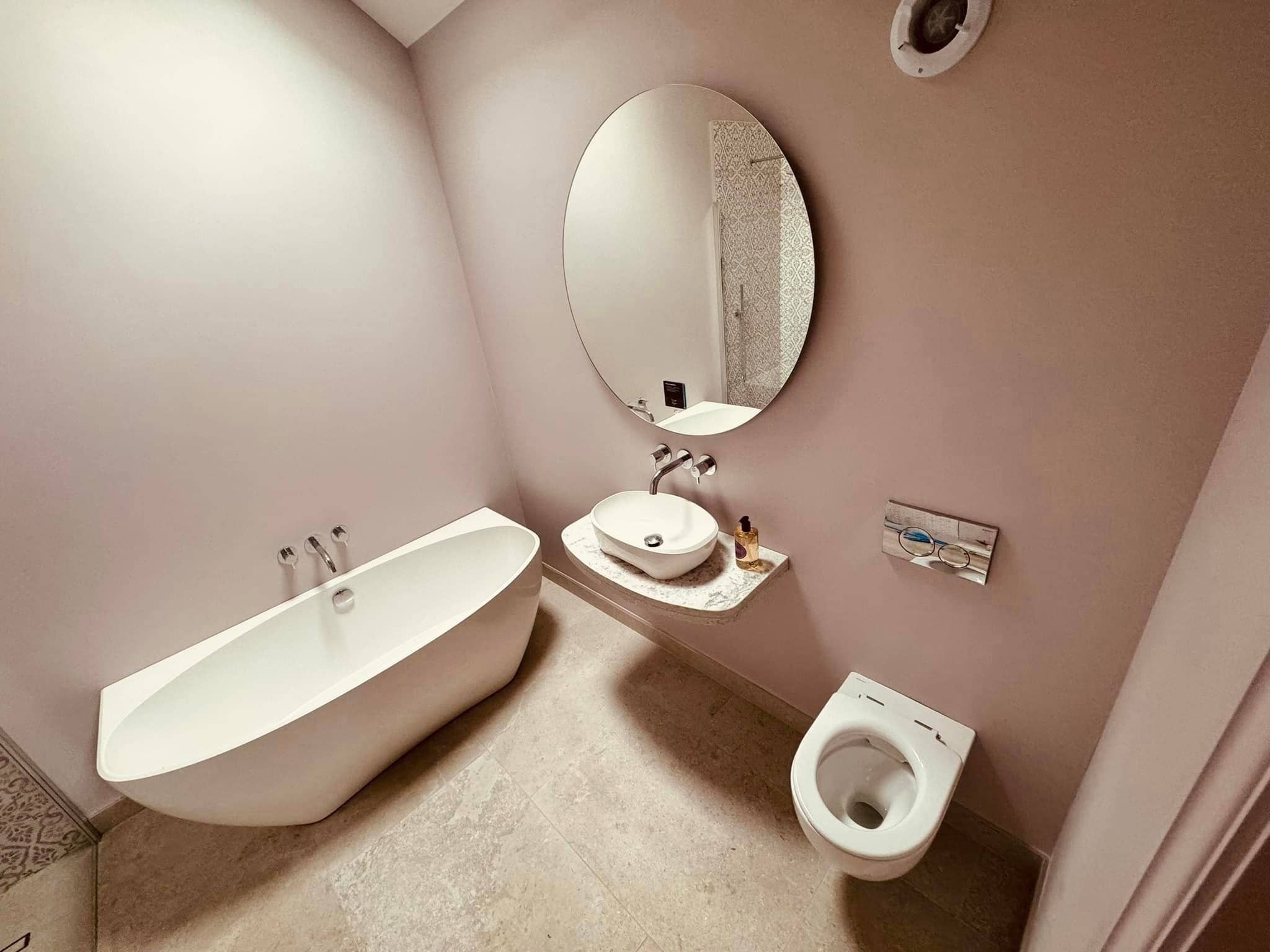 A modern bathroom with a white freestanding bathtub, a round mirror above a vessel sink on a countertop, and a wall-mounted toilet on beige tiled flooring. The walls are painted a soft neutral color.
