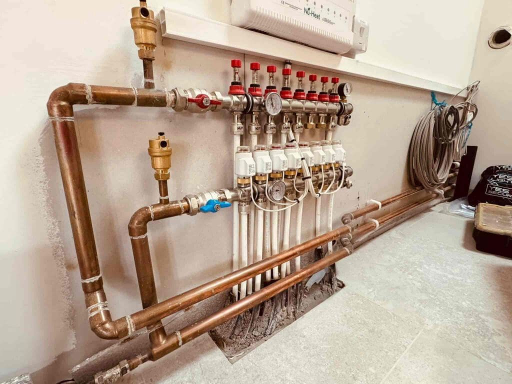 A manifold system with multiple valves, gauges, and copper pipes is installed against a white wall on a tiled floor. Coiled hoses and tools are visible nearby, suggesting ongoing plumbing work.