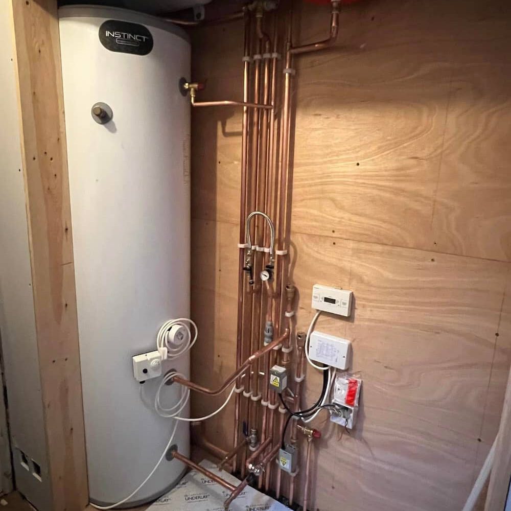 A tall white water heater is mounted on a wall with multiple neatly arranged copper pipes, valves, and electrical controls attached to a plywood wall in a utility room.