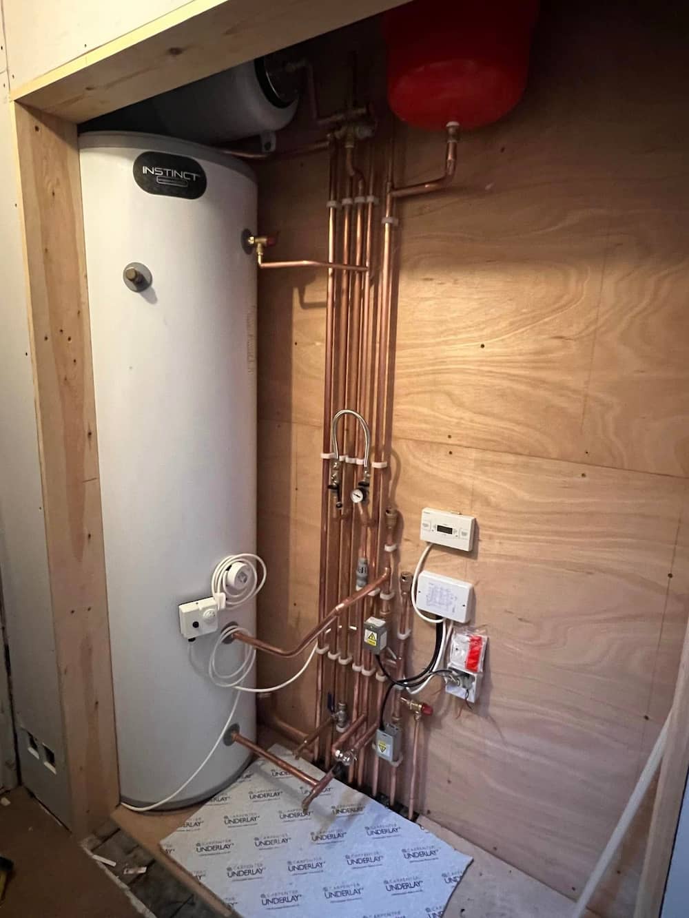A water heater tank is installed in a wooden alcove with neatly arranged copper pipes, a red expansion vessel, electrical outlets, wall-mounted controls, and wiring along the wall.