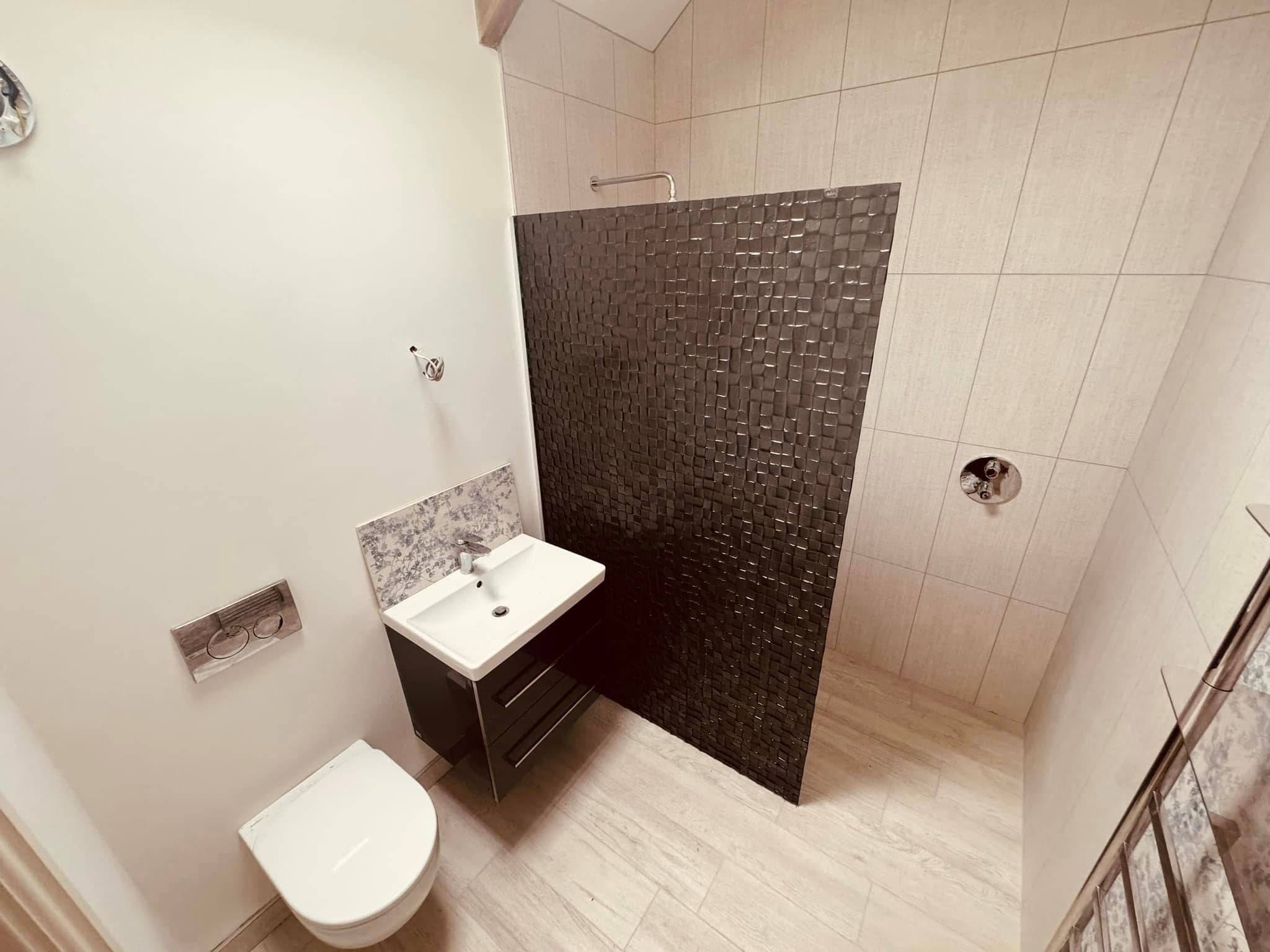Modern bathroom with a wall-mounted toilet, a small white sink on a dark vanity, and a walk-in shower separated by a textured dark partition. The room features light-colored tile walls and floors.