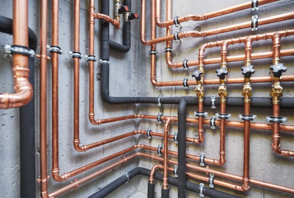 Copper pipes with valves, black insulation, and metal brackets are mounted on a concrete wall, forming a complex plumbing system with multiple bends and connections.