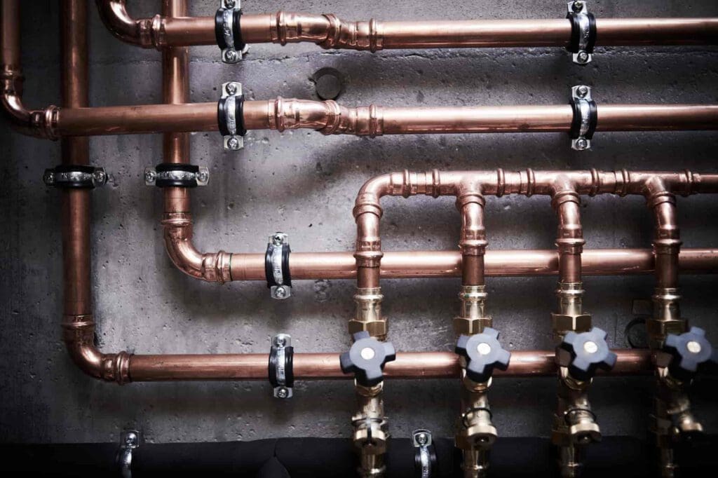 Copper pipes and valves are mounted on a concrete wall, arranged in a neat, organized pattern. The pipes are secured with metal brackets and have black-handled valve controls attached at the bottom.