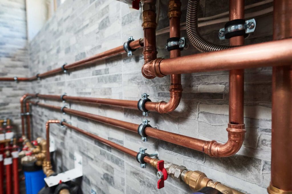 Copper water pipes are mounted along a tiled wall with several valves and connectors. The pipes are secured with metal brackets and run horizontally and vertically in a neat, organized arrangement.