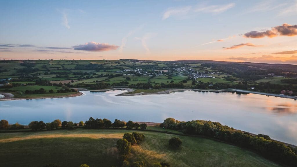 A calm river curves through green fields and rolling hills, with a village in the distance under a colorful, partly cloudy sunset sky. Trees and grassy areas border the water’s edge.