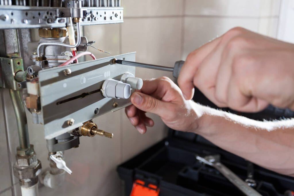 A person uses a screwdriver to repair or adjust a wall-mounted gas boiler, with a toolbox and tools visible in the background.